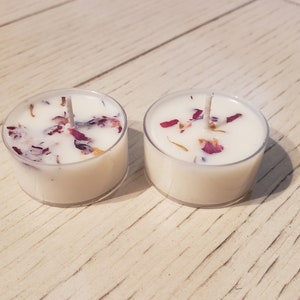 May include: Two white tea light candles with dried flower petals embedded in the wax. The candles are in clear glass containers and are sitting on a light wood surface.