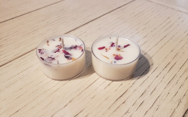 May include: Two white tea light candles with dried flower petals embedded in the wax. The candles are in clear glass containers and are sitting on a light wood surface.