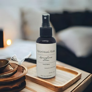 Pu&ograve; includere: Una bottiglia marrone scuro di spray "Spiritual Kids Springtime Linen" con un nebulizzatore nero. L'etichetta recita "Handmade in small batches" e "NET WT. 118 ml". La bottiglia &egrave; su un vassoio di legno.