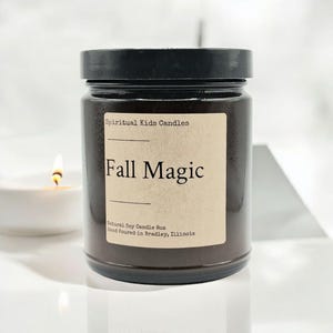 May include: A brown glass candle jar with a black lid. The label is beige with black text that reads "Spiritual Kids Candles" and "Fall Magic". The label also includes the text "Natural Soy Candle 8oz" and "Hand Poured in Bradley, Illinois".