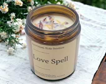 May include: A brown glass jar candle with a beige label that reads "Spiritual Kids Candles Love Spell Natural Soy Candle 4oz Hand Poured in Bradley, Illinois". The candle is topped with a white wax layer and dried flowers.
