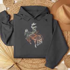 Puede incluir: Una sudadera con capucha gris oscuro con un estampado gráfico de un caballo y un jinete estilizados. El caballo y el jinete están delineados en negro y rellenos con un patrón geométrico en tonos marrones, naranjas y blancos. El texto "HEART POINTED WEST" está impreso debajo del caballo y el jinete.