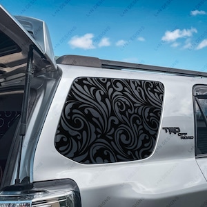 Puede incluir: Un SUV plateado con una calcomanía de ventana de patrón de remolino en blanco y negro. La calcomanía está en la ventana trasera del lado del pasajero. El vehículo tiene un emblema "TRD Off Road" en la parte trasera.