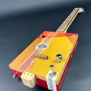 Puede incluir: Una guitarra de caja de puros con un diseño geométrico rojo, amarillo y naranja. La guitarra tiene un mástil de madera y una sola pastilla. La caja está etiquetada "Arturo Fuente"