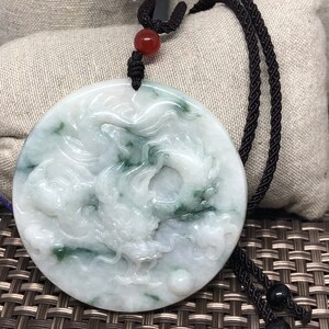 May include: A round jade pendant with a carved dragon design. The pendant is white with green accents and is hanging from a black cord with a red bead.