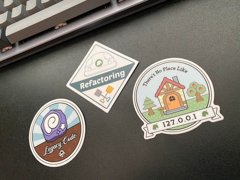 May include: Three stickers featuring a house, a construction worker, and a purple gem. The house sticker has the text "There's No Place Like 127.0.0.1" and a green leaf icon. The construction worker sticker has the text "Refactoring" and a shovel. The gem sticker has the text "Legacy Code" and a skull and crossbones.