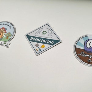 May include: Three round stickers with different designs. The first sticker is blue and green with a house and the text "There's No Place Like 127.0.0.1". The second sticker is a diamond shape with a construction helmet and the text "Refactoring". The third sticker is brown and blue with a spiral and the text "Legacy Code".