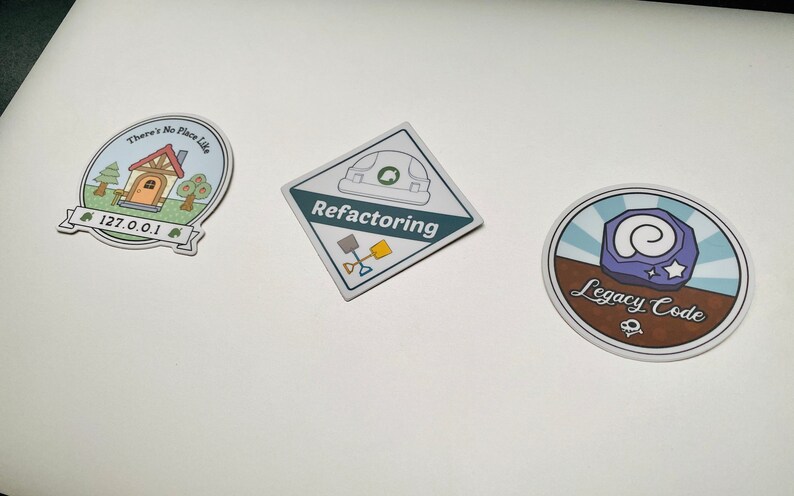 May include: Three round stickers with different designs. The first sticker is blue and green with a house and the text "There's No Place Like 127.0.0.1". The second sticker is a diamond shape with a construction helmet and the text "Refactoring". The third sticker is brown and blue with a spiral and the text "Legacy Code".