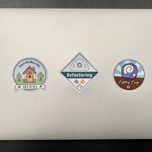 May include: Three stickers on a silver laptop. The left sticker is a blue circle with a house, trees, and the text "There's No Place Like 127.0.0.1". The middle sticker is a diamond shape with the text "Refactoring" and a green construction helmet. The right sticker is a blue circle with a purple swirl and the text "Legacy Code".