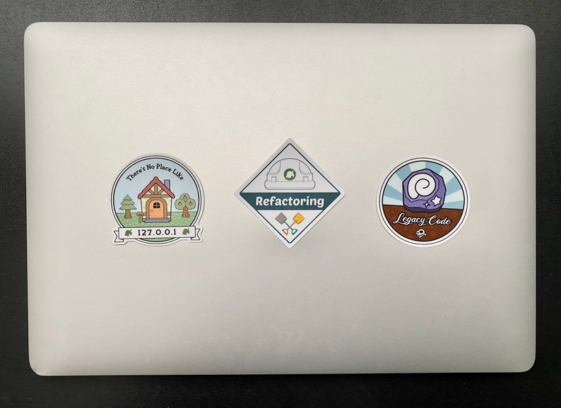 May include: Three stickers on a silver laptop. The left sticker is a blue circle with a house, trees, and the text "There's No Place Like 127.0.0.1". The middle sticker is a diamond shape with the text "Refactoring" and a green construction helmet. The right sticker is a blue circle with a purple swirl and the text "Legacy Code".