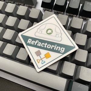 May include: A white sticker with a green and blue design. The sticker features a construction worker's helmet, two shovels, and the text "Refactoring". The sticker is placed on a gray keyboard.