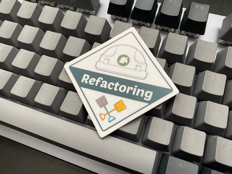 May include: A white sticker with a green and blue design. The sticker features a construction worker's helmet, two shovels, and the text "Refactoring". The sticker is placed on a gray keyboard.