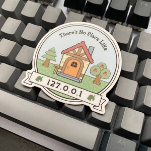 May include: A round sticker with a cartoon house, trees, and the text "There's No Place Like 127.0.0.1".