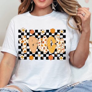 May include: White t-shirt featuring the word "TEACH" in a retro font, with fall-themed designs. The letters are decorated with pumpkins, leaves, and polka dots. The word is set against a black and white checkered background, with floral accents.