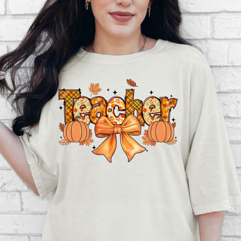 May include: Cream-colored t-shirt with the word "Teacher" in a fall-themed design. The word is decorated with plaid patterns, pumpkins, and autumn leaves. An orange bow and pumpkins are below the word.