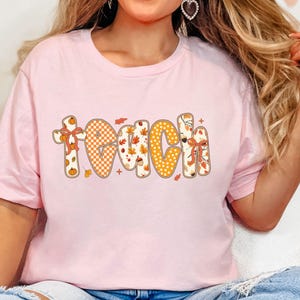 May include: A light pink t-shirt with the word "TEACH" in a fall-themed design. The letters are decorated with pumpkins, leaves, and a checkered pattern in orange and white. The shirt is short-sleeved.