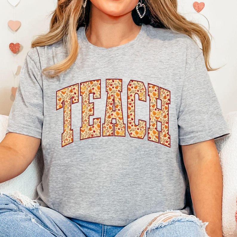 May include: A gray t-shirt with the word "TEACH" in large, block letters across the front. The letters are filled with a fall-themed pattern of leaves, pumpkins, and other autumnal elements. The shirt is a crew neck style.