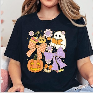 May include: Black t-shirt featuring a whimsical Halloween design. The graphic includes a smiling pumpkin, a reading ghost, cherries with sunglasses, and decorative bows. The design incorporates orange, purple, and yellow colors.
