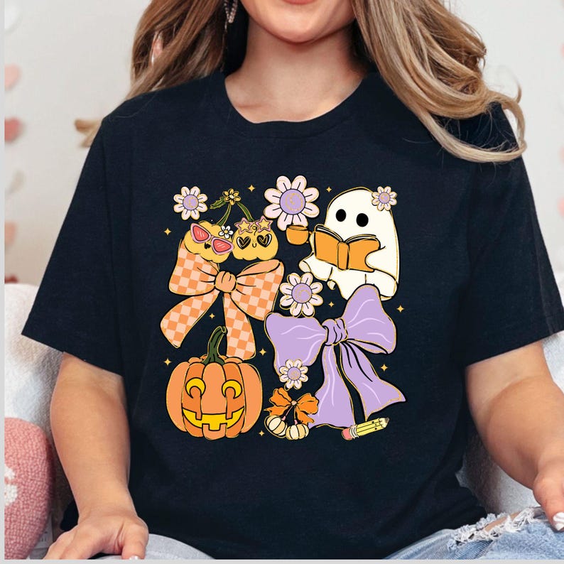 May include: Black t-shirt featuring a whimsical Halloween design. The graphic includes a smiling pumpkin, a reading ghost, cherries with sunglasses, and decorative bows. The design incorporates orange, purple, and yellow colors.