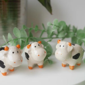 May include: Three ceramic cow figurines with black and white spots and orange hooves. The cows are standing on a white surface.