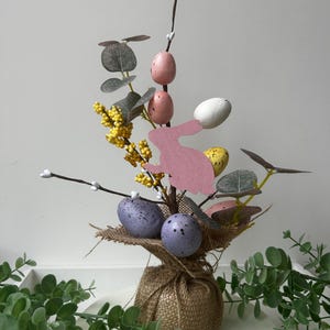 May include: An Easter decoration with speckled eggs in purple, yellow, and white, pink eggs, and a pink bunny cutout. The arrangement is set in a burlap-wrapped base with faux greenery and yellow floral accents, perfect for the spring season.
