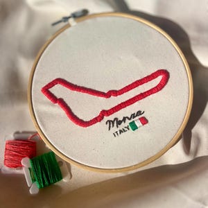 Puede incluir: Un bastidor de bordado circular de madera que contiene tela de color blanco roto con el contorno del circuito de Monza bordado en rojo. El texto Monza ITALY está bordado en hilo negro junto a un pequeño icono de la bandera italiana verde, blanca y roja.