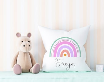 Pode incluir: Uma almofada branca com um desenho de arco-íris e o nome "Freya" impresso em itálico. O arco-íris é composto por listras rosa, roxas, laranja e vermelhas com um pequeno coração laranja no centro. A almofada está sobre uma superfície verde claro com um hipopótamo de pelúcia no fundo.