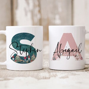 May include: Two white ceramic mugs with personalized designs. One mug features a teal letter "S" with the name "Stephen" and a winter scene. The other mug has a pink letter "A" with the name "Abigail" and a winter scene.