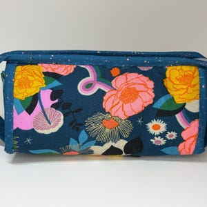 May include: A rectangular, zippered pouch with a floral pattern. The fabric features large pink and yellow flowers, rainbows, and daisies on a dark blue background. The pouch has a dark blue trim and handles.