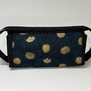 May include: A rectangular fabric pouch featuring a dark blue and black floral pattern with gold-colored centers. The top and side panels are made of black fabric with a small gold cross pattern. It includes a zipper closure with a metallic pull.