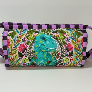 May include: A rectangular cosmetic bag with a teal dinosaur design on a floral background. The bag has a purple and black checkered trim and handles. The zipper is silver.