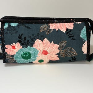 May include: A rectangular floral print bag with a black zipper and handles. The bag features a teal background with large pink and teal flowers. The edges are trimmed with black fabric with white speckles. A silver-colored ring is attached to the zipper.