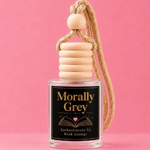 May include: A clear glass car air freshener bottle with a light wood cap and a beige rope. The black label features gold text reading Morally Grey, a graphic of an open book with a small pink heart, and the text Enchantments Co Book Lounge.
