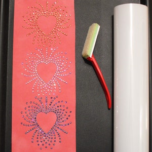 May include: Three heart-shaped designs made of rhinestones on a red background. A white roller and a red squeegee are on a black surface.