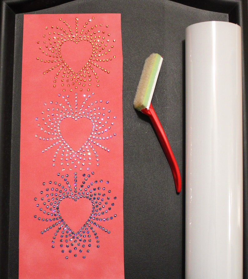 May include: Three heart-shaped designs made of rhinestones on a red background. A white roller and a red squeegee are on a black surface.