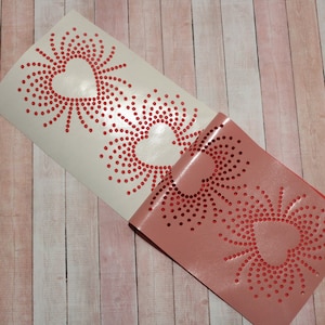 May include: Two white vinyl stencils with a red dot pattern in the shape of a heart. The stencils are on a wooden surface.