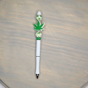 May include: A white pen with a green marijuana leaf design on the top. The pen has green and white beads with a marijuana leaf design.