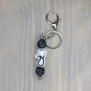 May include: A black and white stethoscope charm keychain with a silver key ring. The charm features a white stethoscope with black accents and black beads with white stethoscope designs.