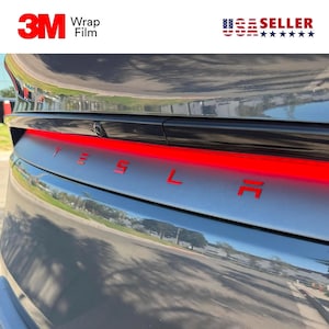 May include: A close-up of the rear of a gray Tesla car with the red letters spelling out "TESLA". The car has a red light bar across the back. The top left corner has the text "3M Wrap Film" in red and white. The top right corner has the text "USA SELLER".