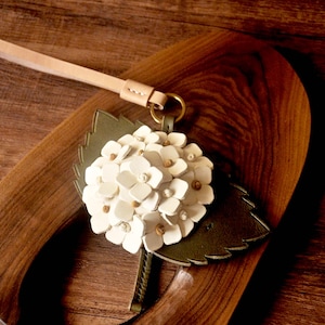 May include: A leather keychain with a white hydrangea flower design and a green leaf backing. The keychain has a tan wrist strap and a gold-coloured ring. The flower has small, tan accents in the centre of each petal.