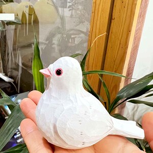 以下が含まれることがあります： 黒い目とピンクのくちばしを持つ白い木製の鳥の置物。鳥は人の手に乗っています。