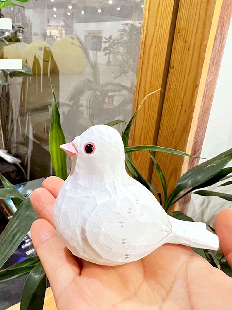 以下が含まれることがあります： 黒い目とピンクのくちばしを持つ白い木製の鳥の置物。鳥は人の手に乗っています。