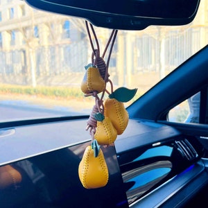 May include: A yellow leather pear keychain hanging from the rearview mirror of a car. The keychain features three pears, each with a stitched design and a green leaf.