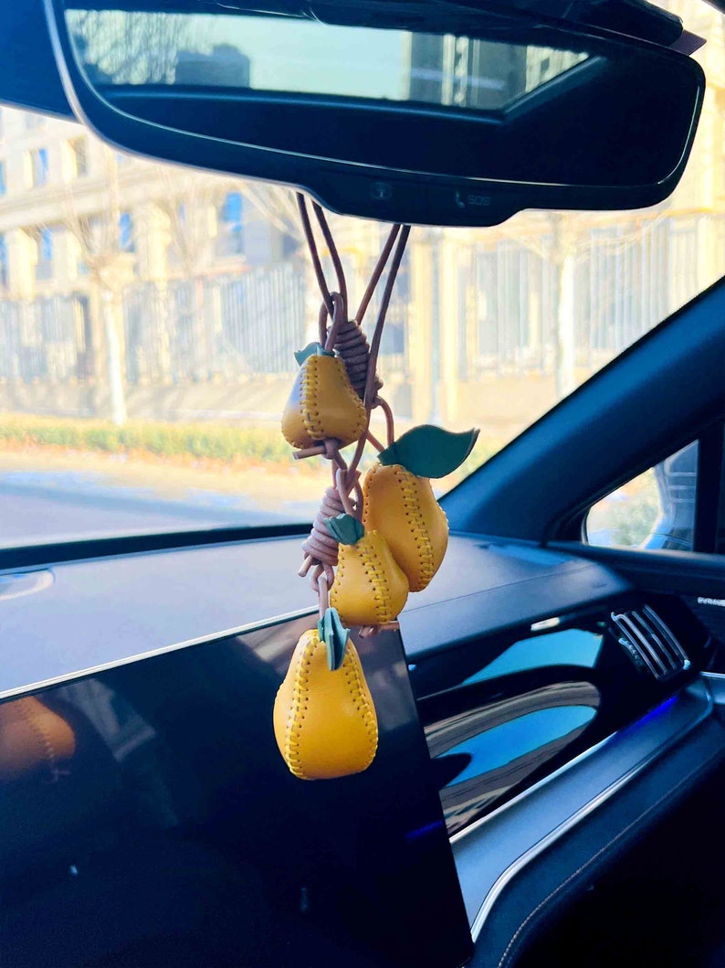 May include: A yellow leather pear keychain hanging from the rearview mirror of a car. The keychain features three pears, each with a stitched design and a green leaf.