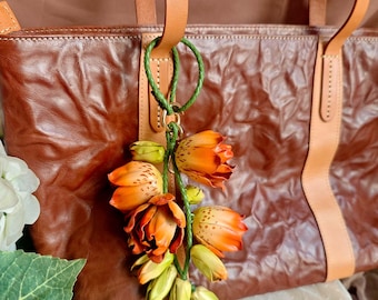 Puede incluir: Un bolso tote de cuero marrón con correas color canela y un llavero trenzado verde. El llavero sostiene un racimo de flores artificiales naranjas y amarillas. La bolsa tiene una apariencia texturizada.