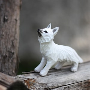 Puede incluir: Una figurita de zorro de madera blanca con ojos negros, de pie sobre una superficie de madera desgastada. El zorro está mirando hacia arriba, con la cabeza inclinada hacia atrás.