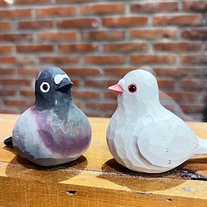 以下が含まれることがあります： 木製の鳥の置物2つ。1つは灰色で黒い目、もう1つは白で赤い目。鳥たちは向き合って、木製の台の上に置かれている。