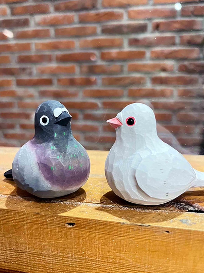 以下が含まれることがあります： 木製の鳥の置物2つ。1つは灰色で黒い目、もう1つは白で赤い目。鳥たちは向き合って、木製の台の上に置かれている。