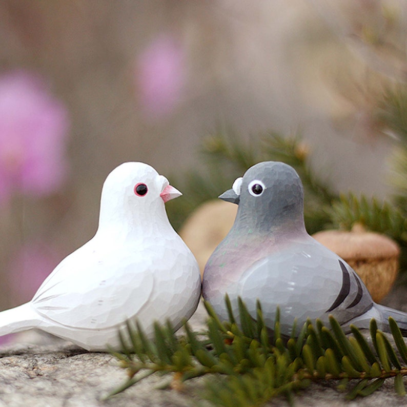 以下が含まれることがあります： 白い木製の鳩の置物と灰色の木製の鳩の置物が、緑色の松葉の上に置かれています。鳩は互いに向き合っており、頭が少し傾いています。白い鳩は赤い目をしており、灰色の鳩は黒い目をしています。