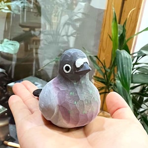 以下が含まれることがあります： グレーとパープルの塗装が施された、手彫りの木製鳥の置物。鳥は白い嘴と白い目を1つ持っています。